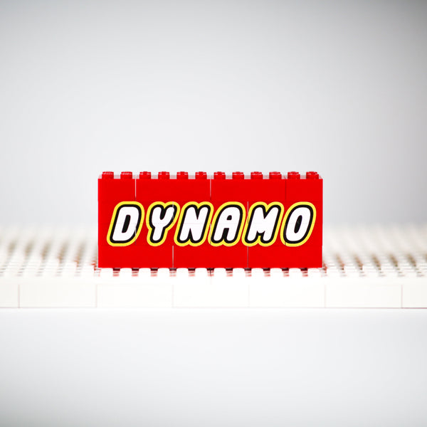 A red toy brick with the word 'DYNAMO' in bold white letters outlined in yellow, standing upright on a white base plate against a light gray studio background.