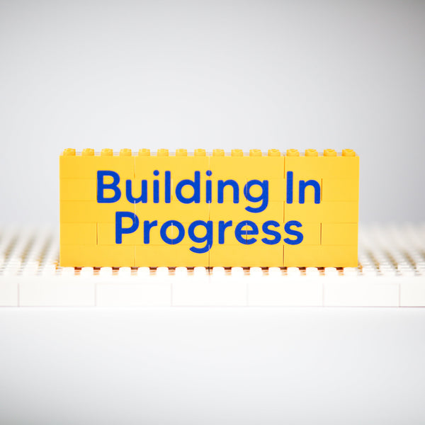 A yellow toy brick block sign with the words 'Building In Progress' in blue, displayed on a white base against a light gray background.