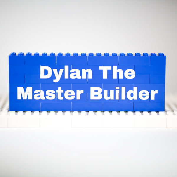 A blue rectangular sign made of toy building blocks stands on a white base plate against a light gray studio background. The sign reads 'Dylan The Master Builder' in white text.