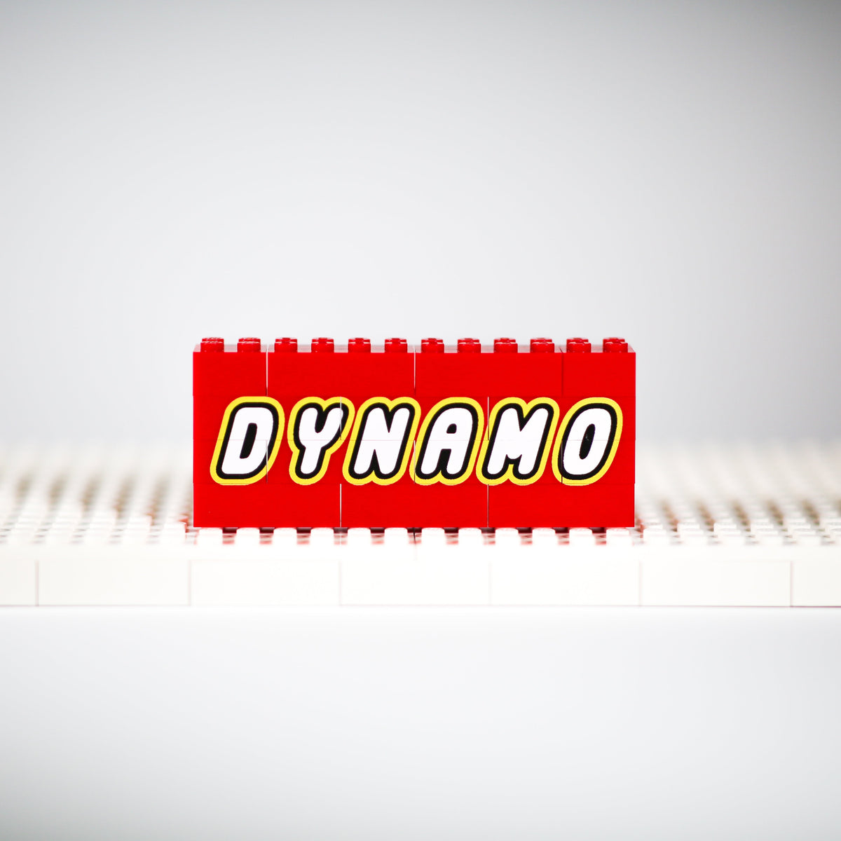 A red toy brick with the word 'DYNAMO' in bold white letters outlined in yellow, standing upright on a white base plate against a light gray studio background.