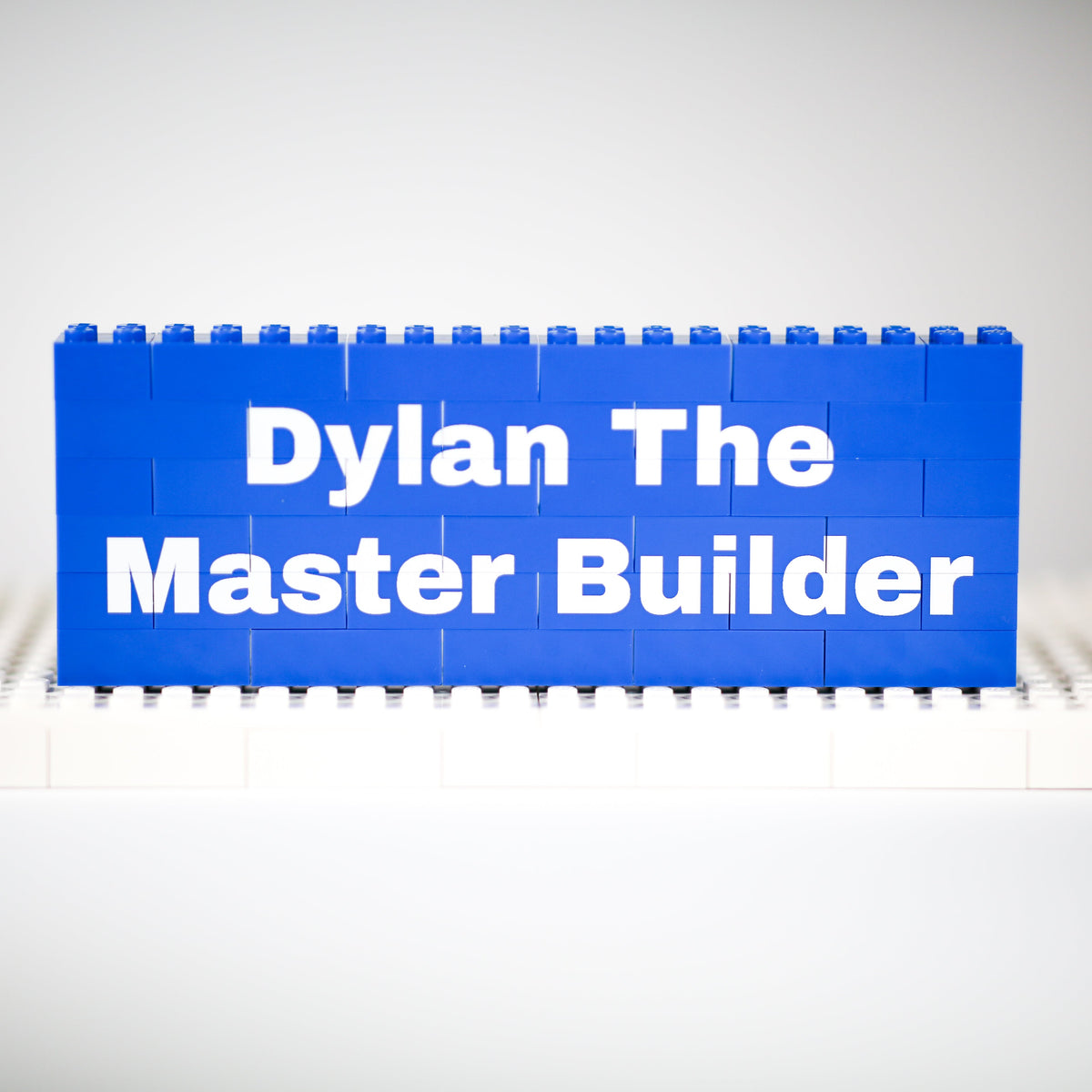 A blue rectangular sign made of toy building blocks stands on a white base plate against a light gray studio background. The sign reads 'Dylan The Master Builder' in white text.